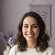 Close-up portrait of a smiling woman with warm skin and dark brown hair, set against a softly blurred dusty lilac and pale ma